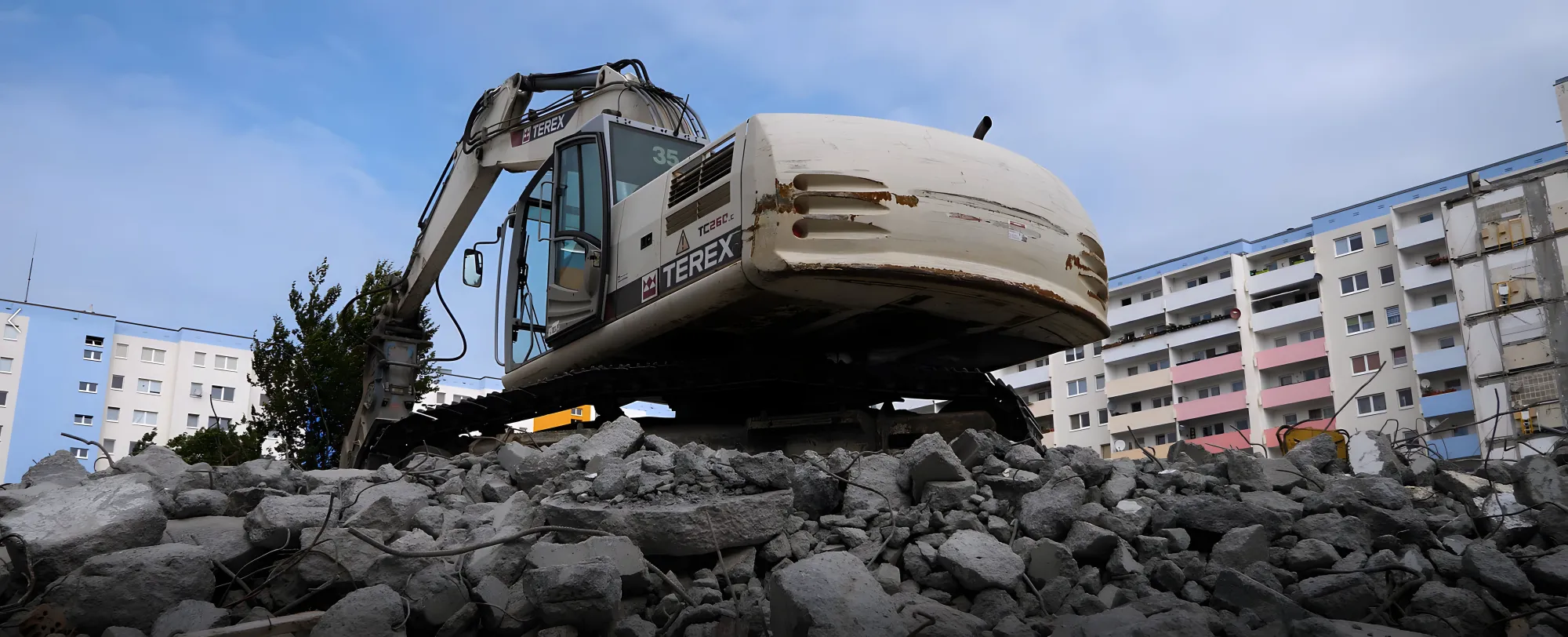 Kettenbagger auf Bauschutt vor farbigen Plattenbauten, Panorama-Luftaufnahme